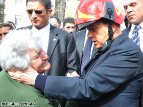 An elderly local resident bursts into tears during a visit by Silvio Berlusconi, wearing a fireman's helmet.
