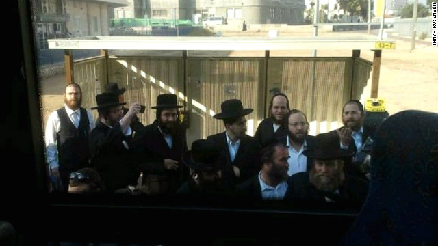 A crowd of ultra-orthodox men gather at a bus stop while Tanya Rosenblit refuses to sit in the back of the bus.