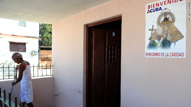 A woman in El Cobre sweeps the entrance to her house, decorated with a poster of the pope.