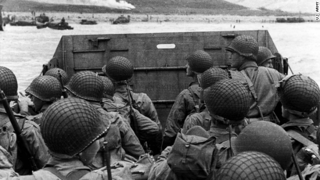 U.S. troops huddle behind the protective front of their landing craft as it nears a beachhead. Smoke in the background is naval gunfire, giving cover to troops on land.