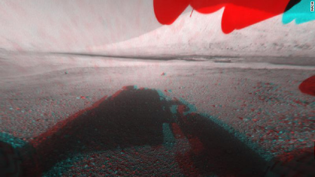 This image is a 3-D view in front of NASA's Curiosity rover. The anaglyph was made from a stereo pair of Hazard-Avoidance Cameras on the front of the rover. Mount Sharp, a peak that is about 3.4 miles high, is visible rising above the terrain, though in one "eye" a box on the rover holding the drill bits obscures the view. 