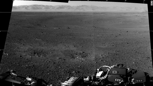 This is a composite image made from two full-resolution images of the Martian surface from the Navigation cameras on the rover, which are located on its "head" or mast. The rim of Gale Crater can be seen in the distance beyond the pebbly ground. The foreground shows two distinct zones of excavation likely carved out by blasts from the rover's descent stage thrusters.