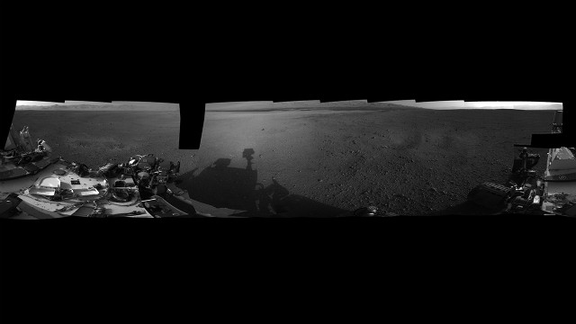 A panoramic photograph shows the Curiosity rover's surroundings at its landing site inside Gale Crater. The rim of Gale Crater can be seen to the left, and the base of Mount Sharp is to the center-right.