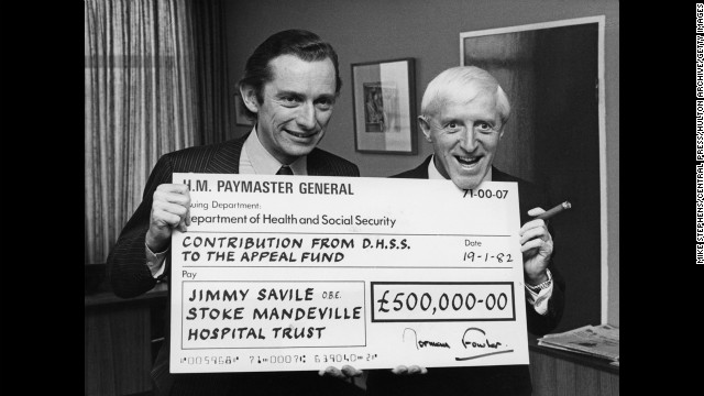 British Secretary of State for Social Services Norman Fowler presents Savile with a check for half a million pounds in 1982 as the government's contribution to an appeal for a new spinal injuries unit at Stoke Mandeville Hospital.