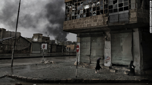 Smoke rises from a fuel station following a mortar attack as Syrian women walk on a rainy day in the Arqub neighborhood of Aleppo on October 25, 2012.