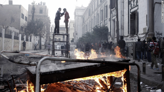 Two protesters stand on top of a piece of furniture while riot police watch from a nearby rooftop in Cairo.