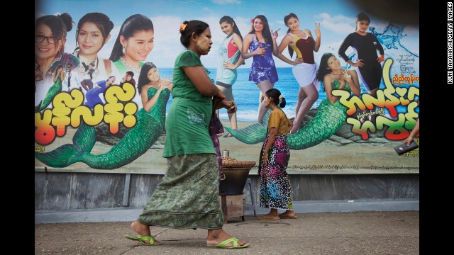 Women wear traditional longyi around their waists in front of a modern movie billboard in Yangon.