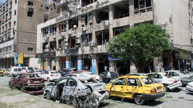 People walk past a damaged building and multiple destroyed cars at the site of an explosion in Damascus where at least 13 were killed on April 30.