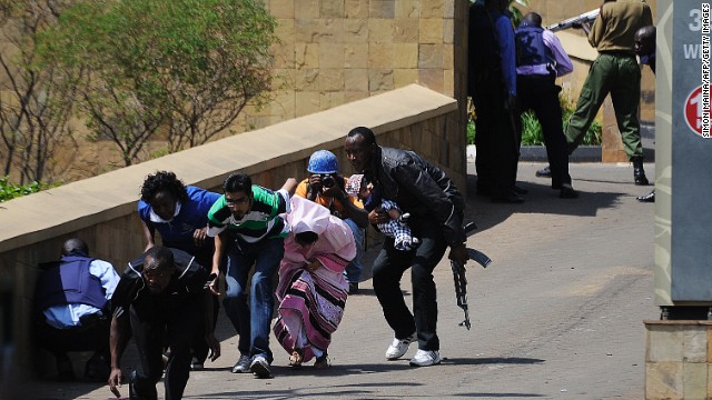 A police officer carries a baby as people keep low and run to safety. Crowds dashed down the streets as soldiers in military fatigues, guns cocked, crawled under cars to get closer to the mall. 