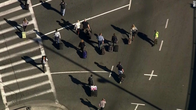 Passengers with luggage cross a street at the airport.
