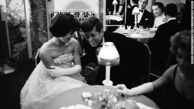 Kennedy lends an ear to his wife as they sit together at a table during cocktail hour before dining at a society gala at the Walford Astoria Hotel in 1960. 