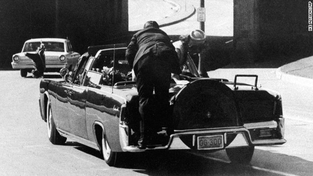 Kennedy slumps in the back seat of the car and his wife leans over to him as Secret Service Agent Clinton Hill rides on the back of the car.