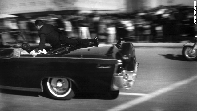 The limousine carrying the mortally wounded President races toward the hospital seconds after three shots are fired. Two bullets hit Kennedy and one hit Connally. Hill rides on the back of the car as the wives cover their stricken husbands.