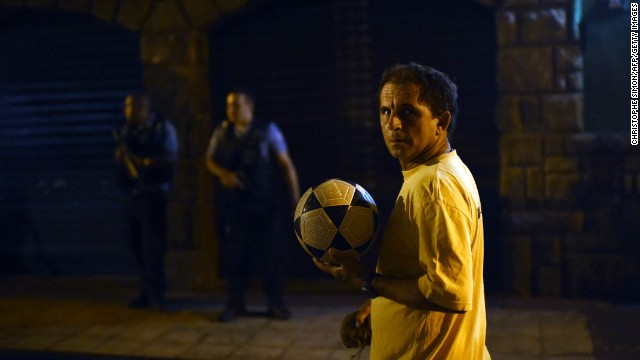 A man walks past with a football in his hand as Brazilian Police Special Forces stand guard in the background during a violent protest in a favela near Copacabana.
