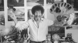 TV painting instructor/artist Bob Ross jubiantly holding up paint pallette & brushes as he stands in front of wall covered w. his landscape paintings in his studio.  (Photo by Acey Harper/The LIFE Images Collection/Getty Images)