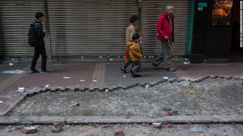 People walk past debris and a section of the pavement which was partial ripped up and used in overnight clashes between protesters and police in the Mongkok area of Hong Kong on February 9, 2016.