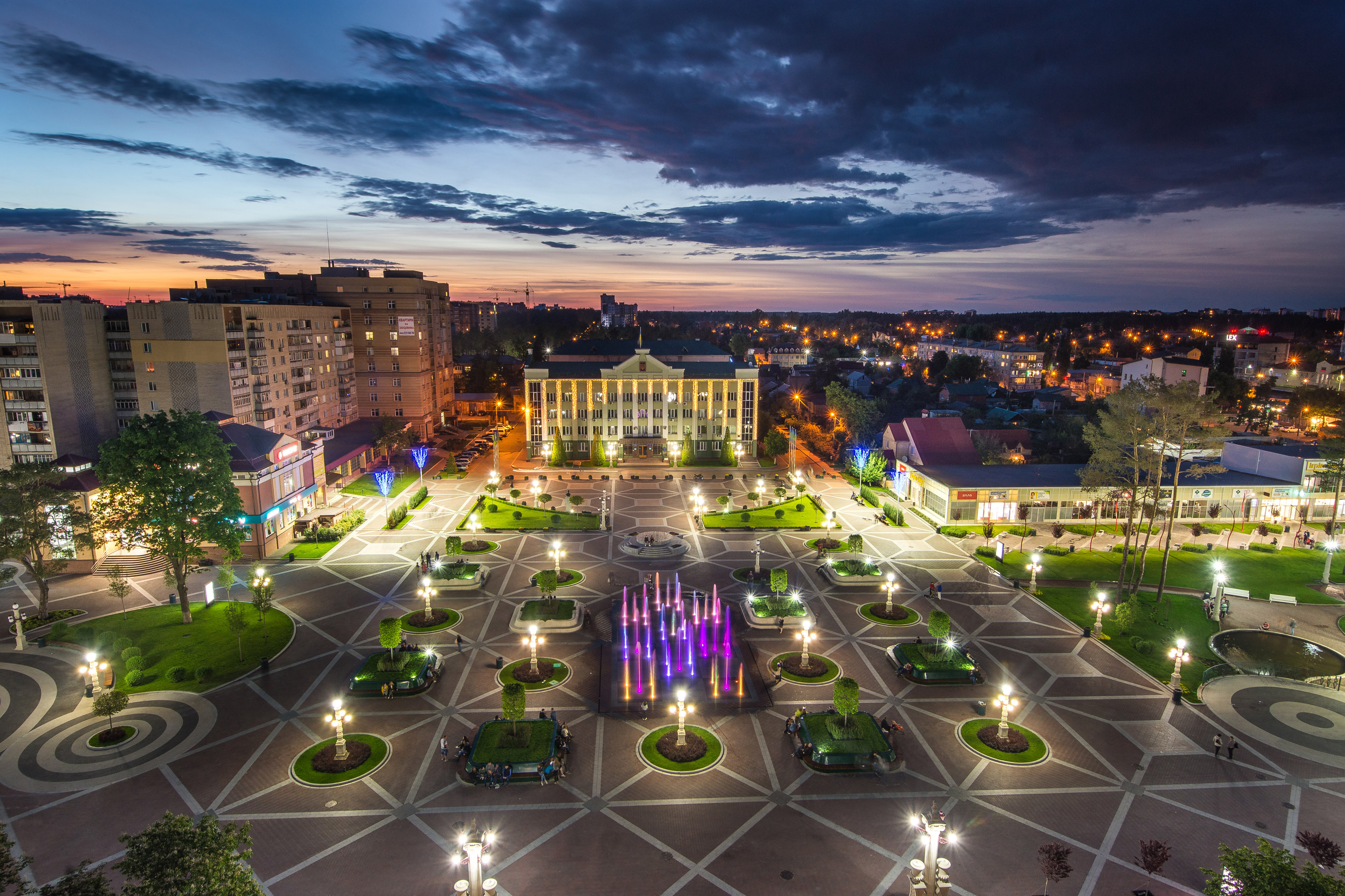 Dusk overlooking a landscaped plaza with green grass areas, ornate lamp posts and a colorful fountain