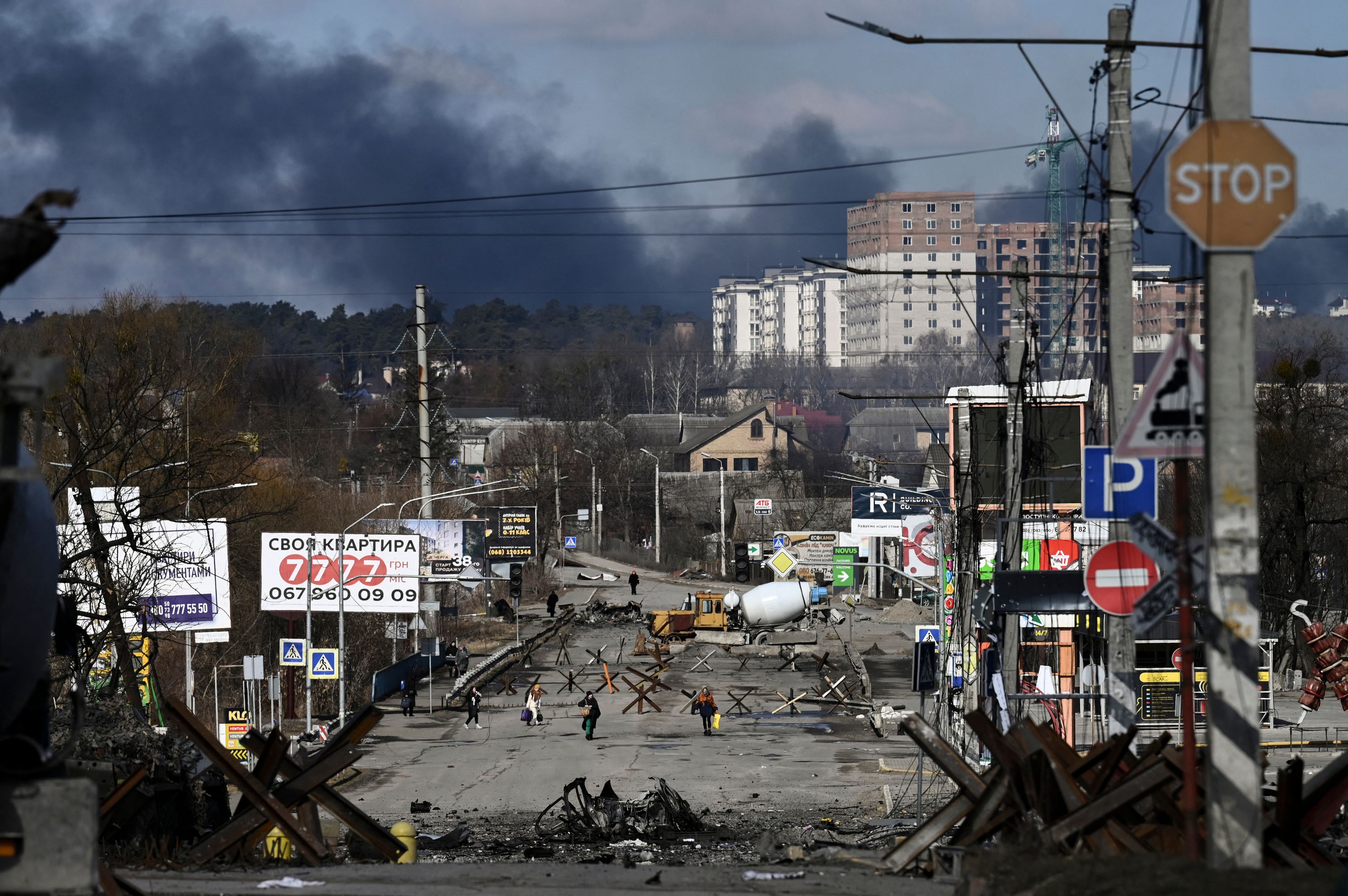 People walking with bags down a road with metal tank traps. The sky is filled with thick grey smoke