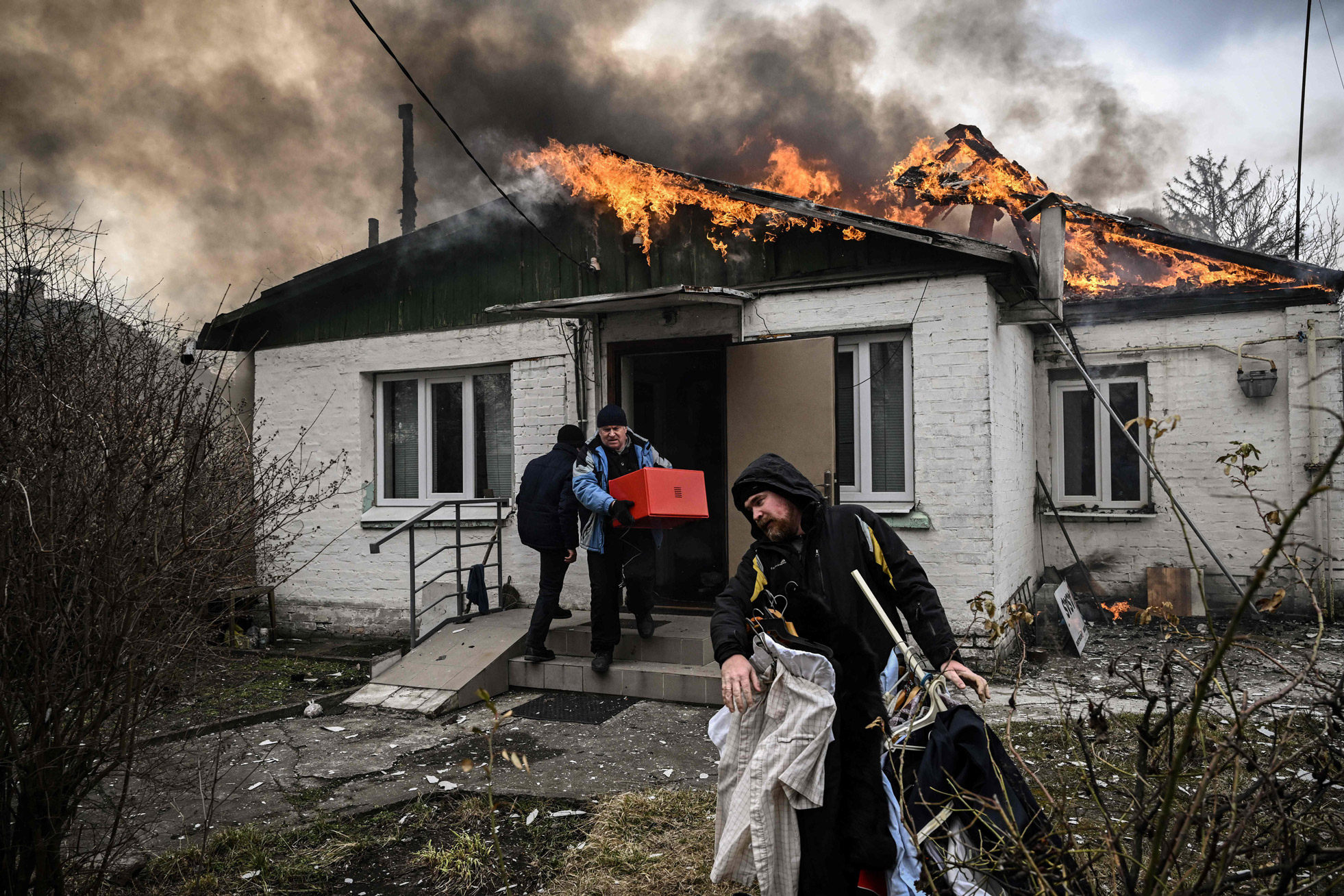 The roof of a house is on fire while two men carry clothes and a box out.