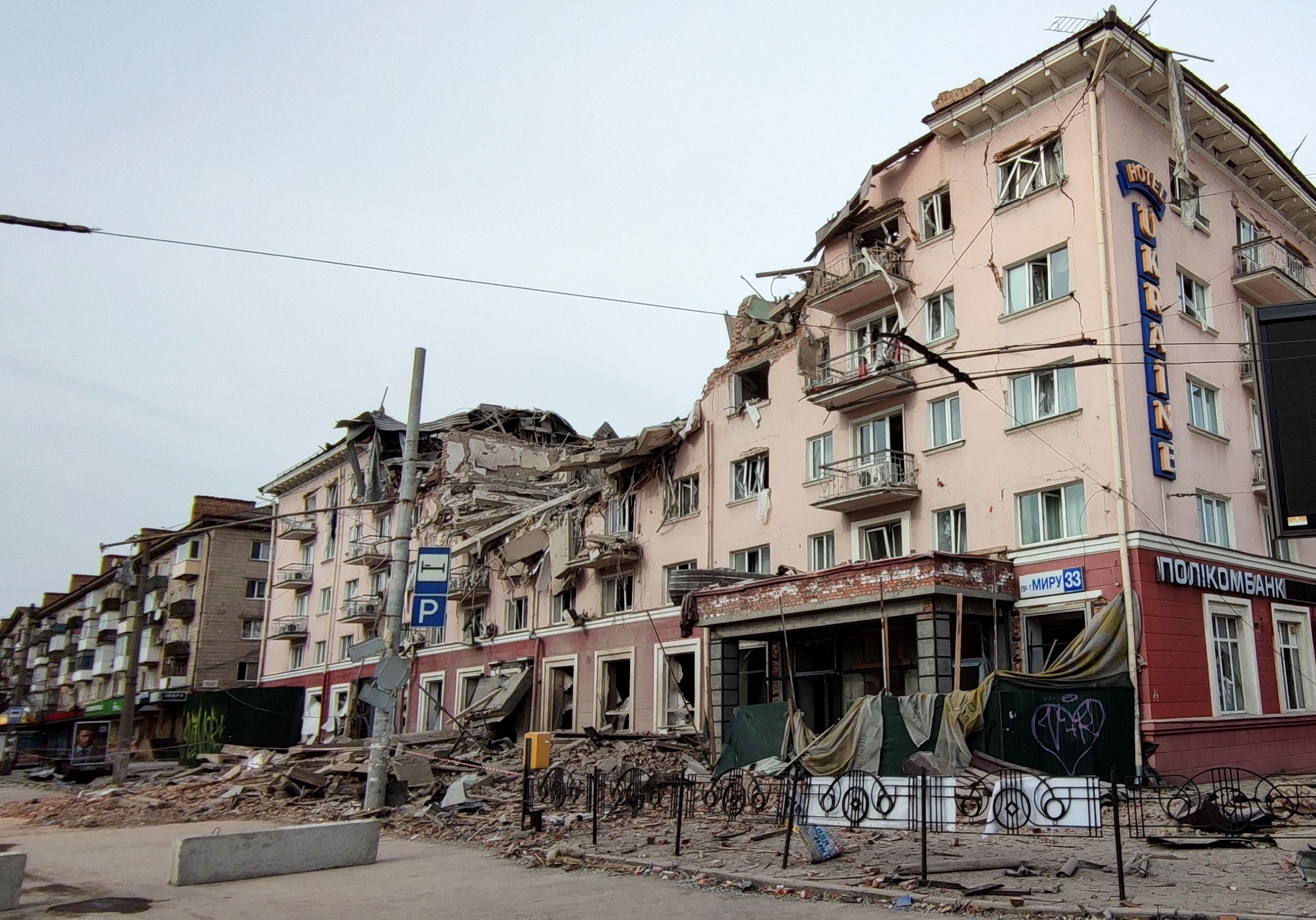 A large building has a huge crater in it, windows are blown out and rubble outside