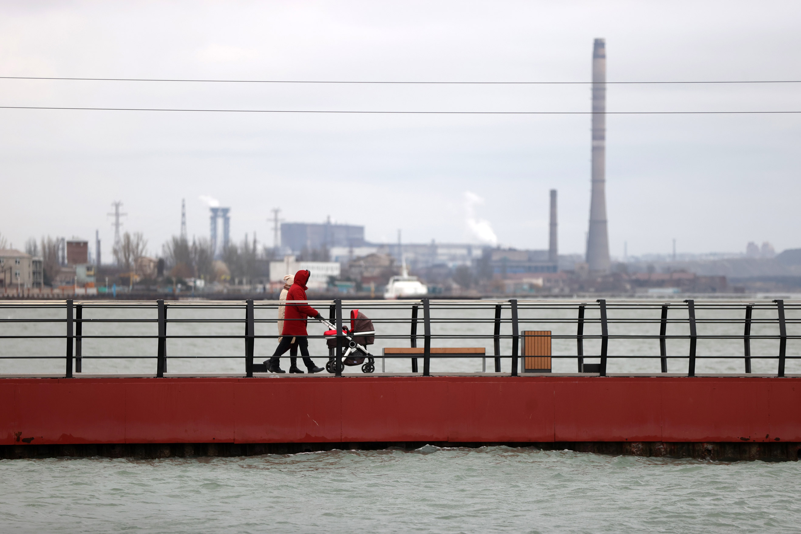 A couple push a pram along a pier with industrial buildings in the background