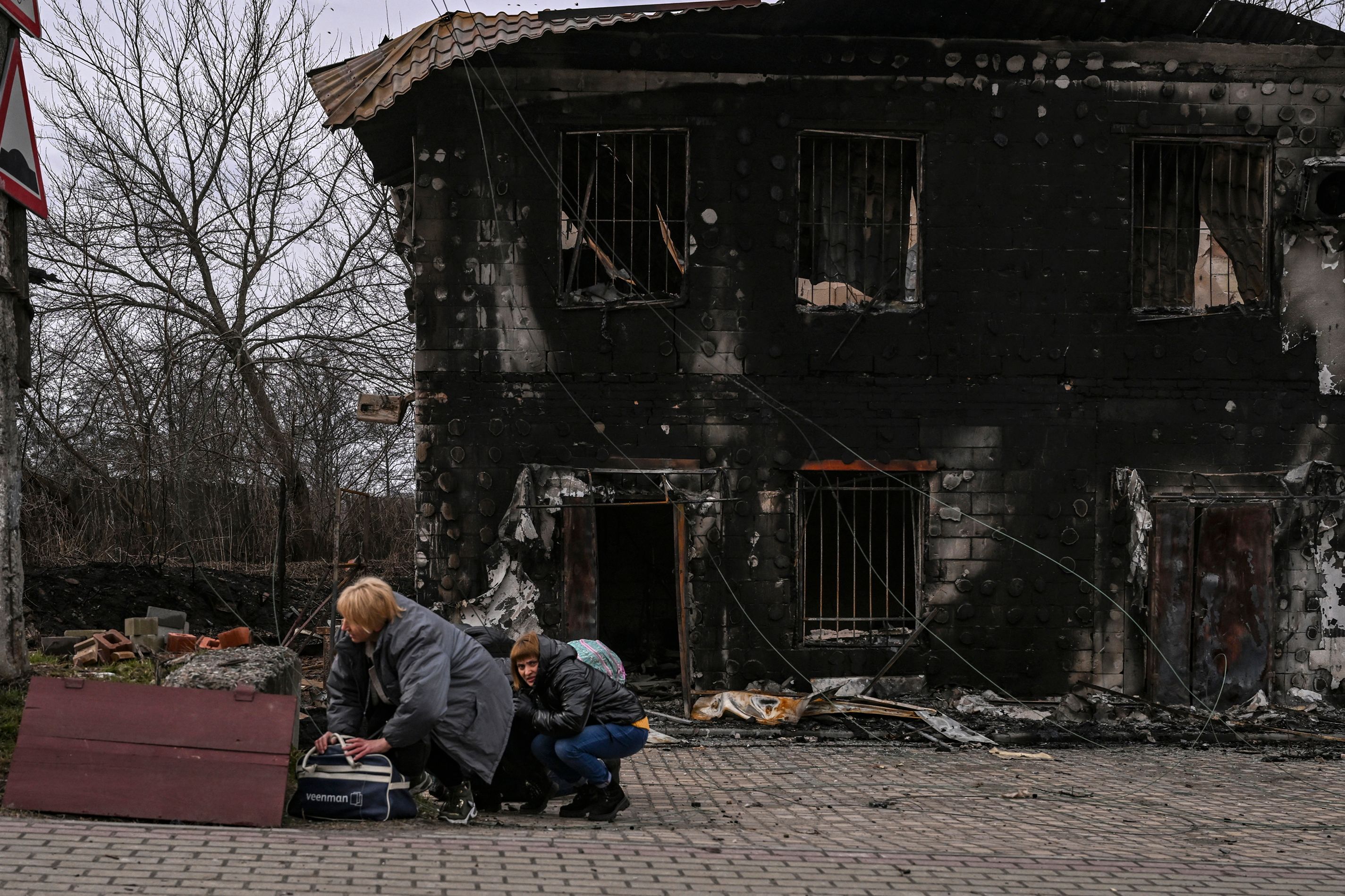 Two women crouching down to take cover outside a burnt-out house surrounded by rubble.